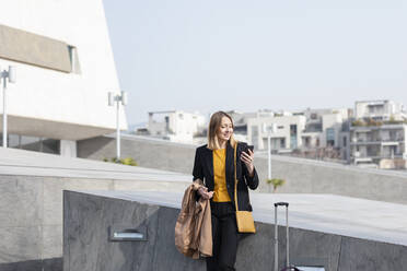 Entrepreneur using mobile phone while standing with jacket by suitcase - EIF00669