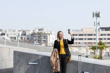 Lächelnde Geschäftsfrau mit Jacke und Koffer, die ein Selfie mit ihrem Mobiltelefon in der Stadt macht - EIF00670