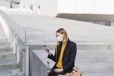 Unternehmerin mit Gesichtsmaske, die ein Mobiltelefon benutzt, während sie an einer Treppe sitzt - EIF00674