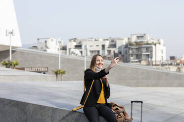 Lächelnde Geschäftsfrau, die ein Selfie mit ihrem Handy macht, während sie auf einer Stützmauer sitzt - EIF00676