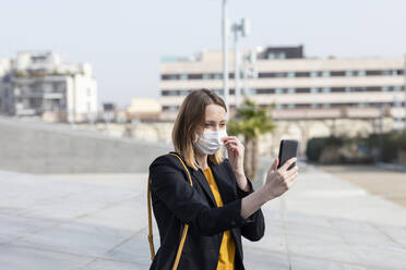 Geschäftsfrau nimmt Gesichtsmaske ab, während sie auf dem Gehweg ein Selfie mit dem Handy macht - EIF00678