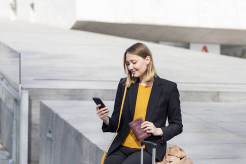 Unternehmer mit Reisepass, der ein Mobiltelefon benutzt, während er auf einer Stützmauer sitzt - EIF00681