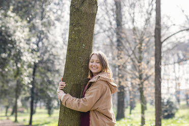 Glückliche erwachsene Frau, die einen Baum umarmt, während sie im Park steht - EIF00698