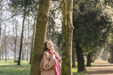 Lächelnde Frau lehnt an einem Baum im Park - EIF00701