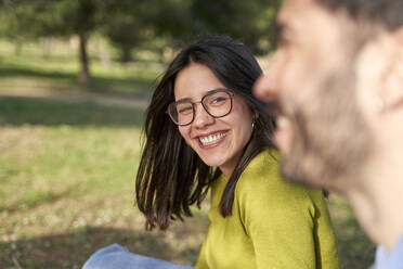 Cheerful woman with eyeglasses looking at boyfriend while having fun in public park - VEGF04080