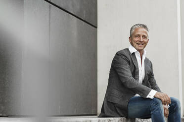 Smiling male restaurant manager looking away while sitting against gray wall - SAJF00109