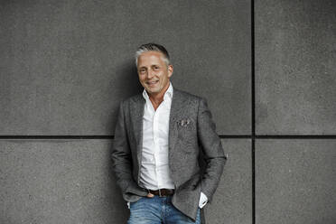 Handsome male entrepreneur with hands in pockets standing against gray wall - SAJF00121