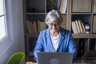 Lächelnde Unternehmerin bei der Arbeit am Laptop vor dem Bücherregal zu Hause - SIPF02280