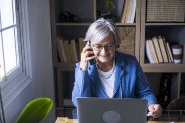 Lächelnde ältere weibliche Fachkraft, die mit einem Mobiltelefon spricht, während sie auf einen Laptop im Heimbüro schaut - SIPF02281
