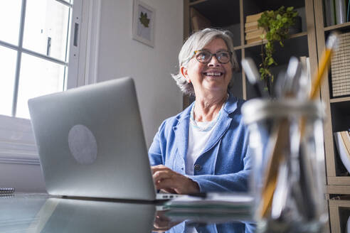 Glückliche Geschäftsfrau, die wegschaut, während sie zu Hause mit ihrem Laptop arbeitet - SIPF02304