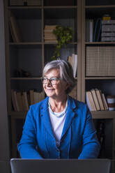 Smiling businesswoman looking away while sitting against bookshelf at home - SIPF02305