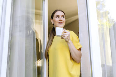 Smiling woman looking away while holding coffee cup at window - OGF00971