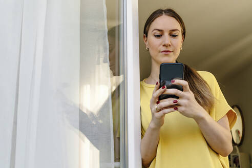 Mid adult woman using smart phone while leaning on window - OGF00975