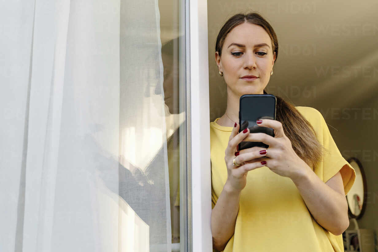 Mid adult woman using smart phone while leaning on window stock photo