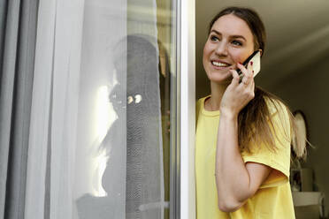 Lächelnde Frau, die wegschaut, während sie am Fenster mit einem Mobiltelefon spricht - OGF00976