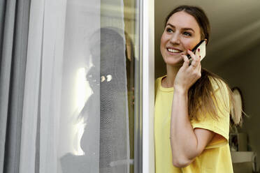Smiling woman looking away while talking on mobile phone at window - OGF00976