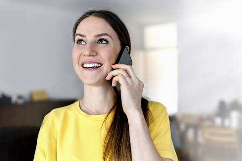 Glückliche Frau mit grauen Augen, die wegschaut, während sie zu Hause mit dem Handy telefoniert - OGF00977