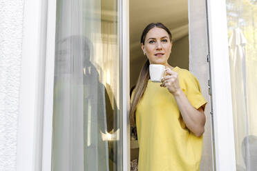 Braunhaarige Frau mit Kaffeetasse am Fenster stehend - OGF00978