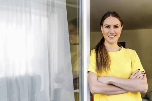 Schöne lächelnde Frau mit verschränkten Armen, die sich zu Hause an ein Fenster lehnt - OGF00979