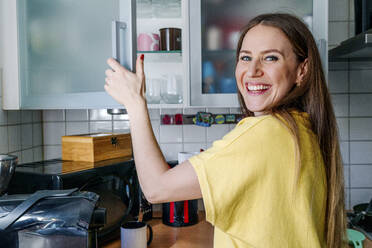 Glückliche Frau mit grauen Augen beim Öffnen einer Schranktür in der Küche zu Hause - OGF00981
