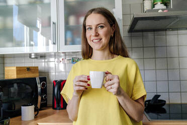 Lächelnde braunhaarige Frau hält Kaffeetasse in Küche zu Hause - OGF00982