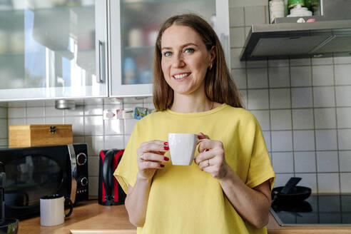 Smiling brown haired woman holding coffee cup in kitchen at home - OGF00982