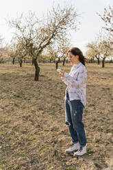Frau zeichnet in Notizblock, während sie an Mandelbäumen steht - MGRF00179