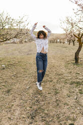 Playful woman jumping with hand raised at almond trees during sunset - MGRF00184