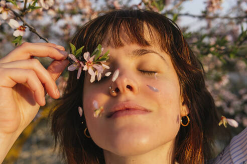 Smiling woman holding almond blossom branch by eyes during sunset - MGRF00198