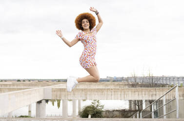 Playful woman in floral dress jumping with hands raised in park - JCCMF01479