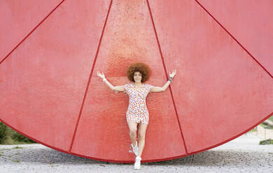 Young woman with arms outstretched leaning on curve red wall - JCCMF01492