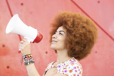 Smiling woman with megaphone by red wall - JCCMF01495