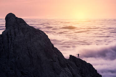 Silhouette einer nicht erkennbaren Person, die ihre Arme ausstreckt und auf dem Gipfel einer rauen Klippe steht, mit bunten Wolken im Hintergrund bei Sonnenuntergang - ADSF22055
