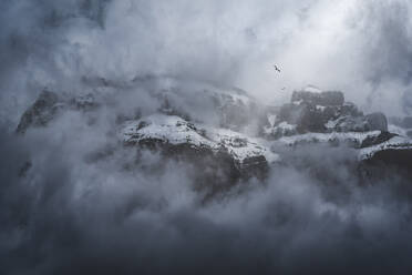 Scharfe, teilweise schneebedeckte Berggipfel, umgeben von dunstigem Nebel unter bewölktem Himmel im Winter - ADSF22070