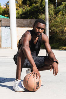 Entschlossener afroamerikanischer Mann in Sportkleidung hockt auf einem Basketballplatz in der Stadt mit einem Ball und schaut in die Kamera - ADSF22092
