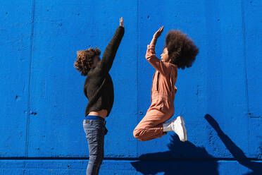 Seitenansicht eines aufgeregten afroamerikanischen lockigen Jungen und eines Mädchens, die Spaß haben und sich gegenseitig High Five geben, während sie den sonnigen Tag auf der Straße vor einer blauen Wand genießen - ADSF22109