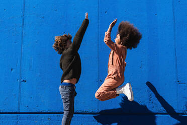 Side view of excited African American curly haired boy and girl having fun and giving high five to each other while enjoying sunny day together on street against blue wall - ADSF22109