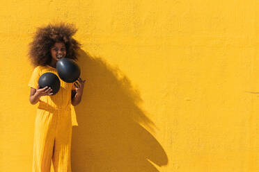 Glückliches afroamerikanisches Teenager-Mädchen mit lockiger Frisur, das einen schwarzen Luftballon hält und vor einem gelben Hintergrund steht - ADSF22112
