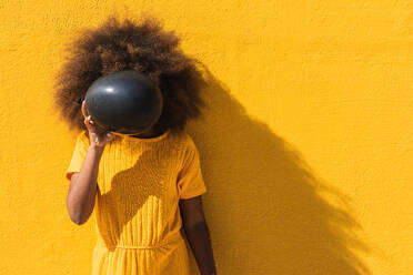 Unrecognizable curly haired teen African American girl in yellow shirt covering face with black balloons while standing against bright yellow wall - ADSF22114