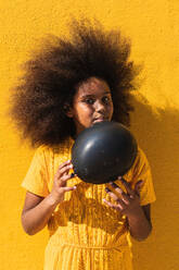 African American teen girl with curly hairstyle holding black balloon while standing against yellow background looking away - ADSF22115