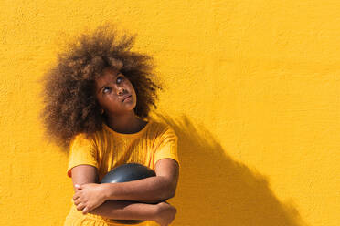 Nachdenkliches afroamerikanisches Teenager-Mädchen mit lockiger Frisur, das einen schwarzen Ballon hält, während es vor einem gelben Hintergrund steht und nach oben schaut - ADSF22116