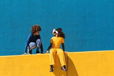 Low angle full body of African American boy communicating with girlfriend wearing VR headset while playing virtual game near colorful wall - ADSF22121