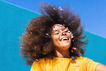 Low angle of happy carefree teen African American girl with long curly hair and colorful confetti on face having fun against blue wall in sunny summer day - ADSF22122