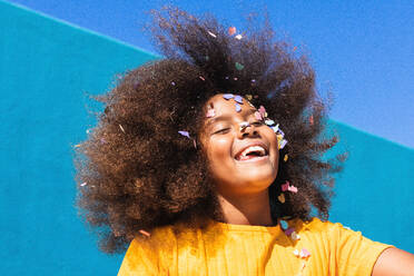 Niedriger Winkel von glücklichen sorglosen Teenager afroamerikanischen Mädchen mit langen lockigen Haaren und bunten Konfetti im Gesicht Spaß gegen blaue Wand in sonnigen Sommertag - ADSF22122