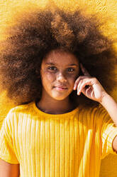 Jugendliches afroamerikanisches Mädchen mit lockigem Haar, das ein gelbes Sweatshirt trägt, die Schläfe berührt und nachdenklich in die Kamera schaut, vor einem gelben Hintergrund - ADSF22131