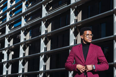 Selbstbewusster junger Afroamerikaner in modischem Anzug und mit Brille steht auf der Straße in der Nähe eines modernen Gebäudes mit Glaswänden an einem sonnigen Tag - ADSF22133