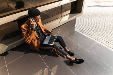 From above of concentrated young African American male freelancer in trendy suit and and sunglasses talking on smartphone and working on laptop while sitting on building stairs on street - ADSF22144