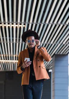 Content African American male in trendy hat and clothes walking along urban street and browsing mobile phone - ADSF22146