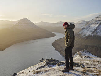 Man on snowy mountain looking towards the ocean in the Faroe Islands - CAVF93738