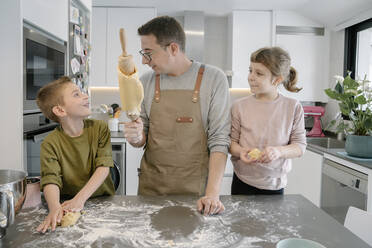 Father showing dough on rolling pin to son while standing by daughter at home - AMPF00141